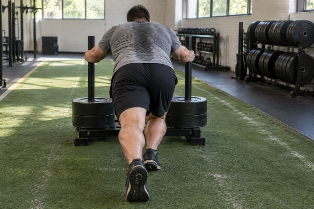 A heavyset man seen from behind, back to the camera, driving a loaded push sled down a turf lane