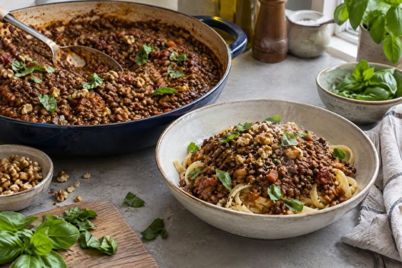A pan of dark lentil and walnut bolognese beside a bowl of pasta with torn basil on top